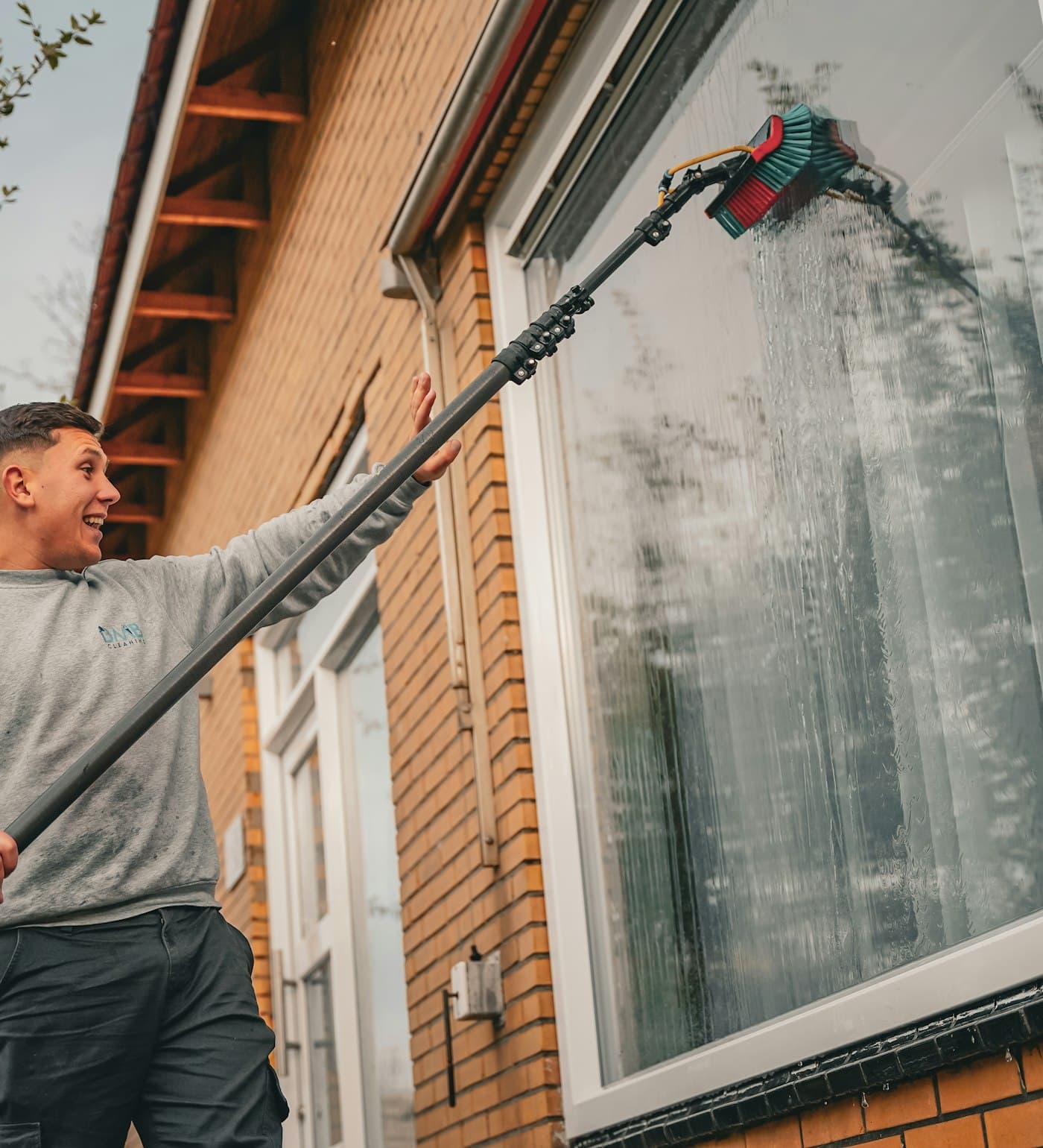 A window cleaner scrubbing the upstairs windows of a brick house with a water-fed pole